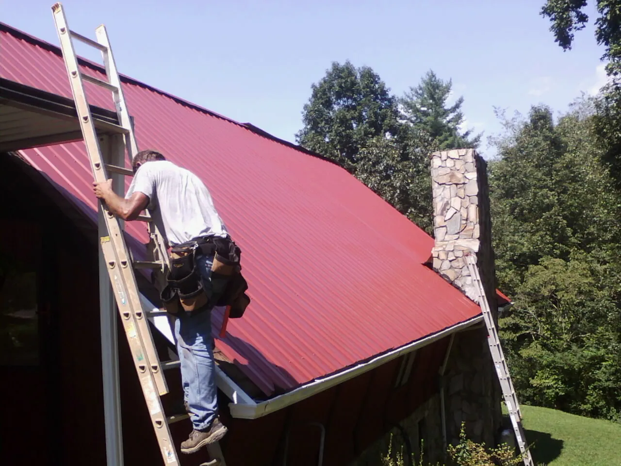 Roofer on a ladder beside a red metal roof and stone chimney — Soffit & Fascia Repair by Roofing Brothers in Congerville